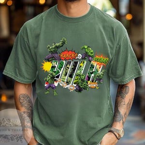 May include: Green t-shirt featuring a comic book style graphic of the Hulk. The design includes the word "Hulk" in a bold font, along with action words like "RAAACK!" and "BOOM!" and images of the Hulk in action.