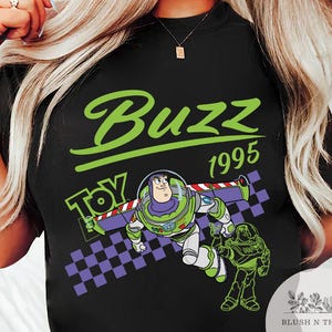 May include: Black t-shirt featuring a graphic of Buzz Lightyear from Toy Story. The design includes the word "Buzz" in green, the year "1995", and the word "Toy". The image also shows a checkered pattern.