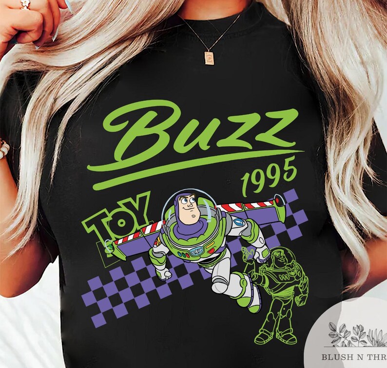 May include: Black t-shirt featuring a graphic of Buzz Lightyear from Toy Story. The design includes the word "Buzz" in green, the year "1995", and the word "Toy". The image also shows a checkered pattern.