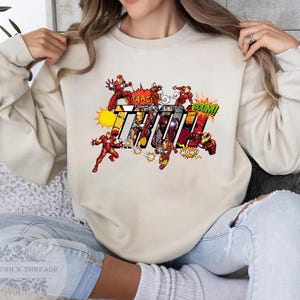 May include: A beige sweatshirt featuring a comic book-style graphic of Iron Man. The design includes Iron Man figures in action poses, with the word "IRON" in bold, colorful letters, and comic book sound effects.