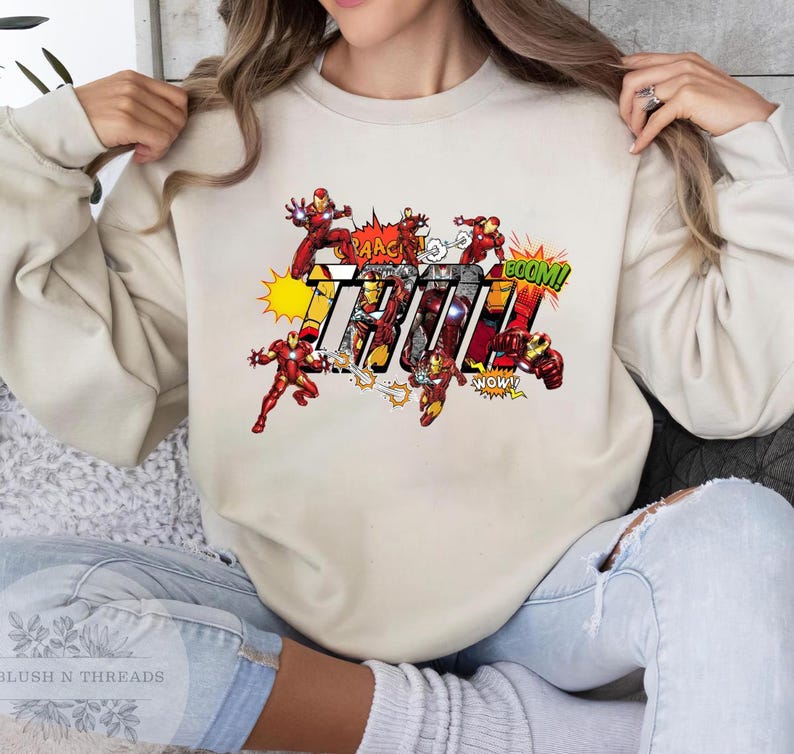 May include: A beige sweatshirt featuring a comic book-style graphic of Iron Man. The design includes Iron Man figures in action poses, with the word "IRON" in bold, colorful letters, and comic book sound effects.