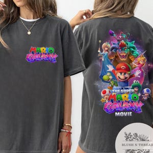 Puede incluir: Camiseta gris oscuro con el logo "Mario Galaxy" en la parte delantera y un gr&aacute;fico colorido de Mario y sus amigos en la parte trasera. La camiseta es de cuello redondo y manga corta.