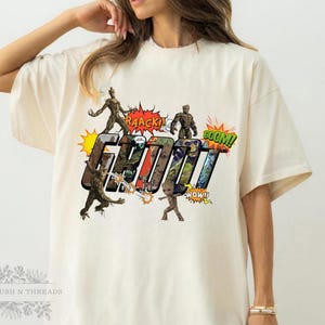 May include: Cream-colored t-shirt featuring a comic-style graphic of the character Groot from Guardians of the Galaxy. The design includes the word "GROOT" in bold letters, surrounded by action bubbles with phrases like "RAACK!" and "BOOM!"