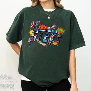 May include: Dark green t-shirt featuring the name "STEVE" in a comic book style with Captain America illustrations. The design includes action words like "CRAACK!" and "BOOM!" in speech bubbles. The shirt has a vintage, worn look.