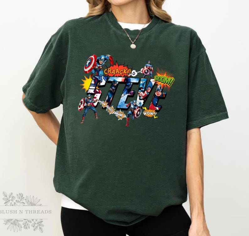 May include: Dark green t-shirt featuring the name "STEVE" in a comic book style with Captain America illustrations. The design includes action words like "CRAACK!" and "BOOM!" in speech bubbles. The shirt has a vintage, worn look.