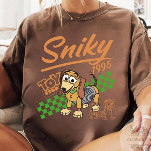 May include: Brown t-shirt featuring the text "Sniky 1995" in orange, with a cartoon image of Slinky Dog from Toy Story. The shirt also includes the Toy Story logo and a green and white checkered pattern.