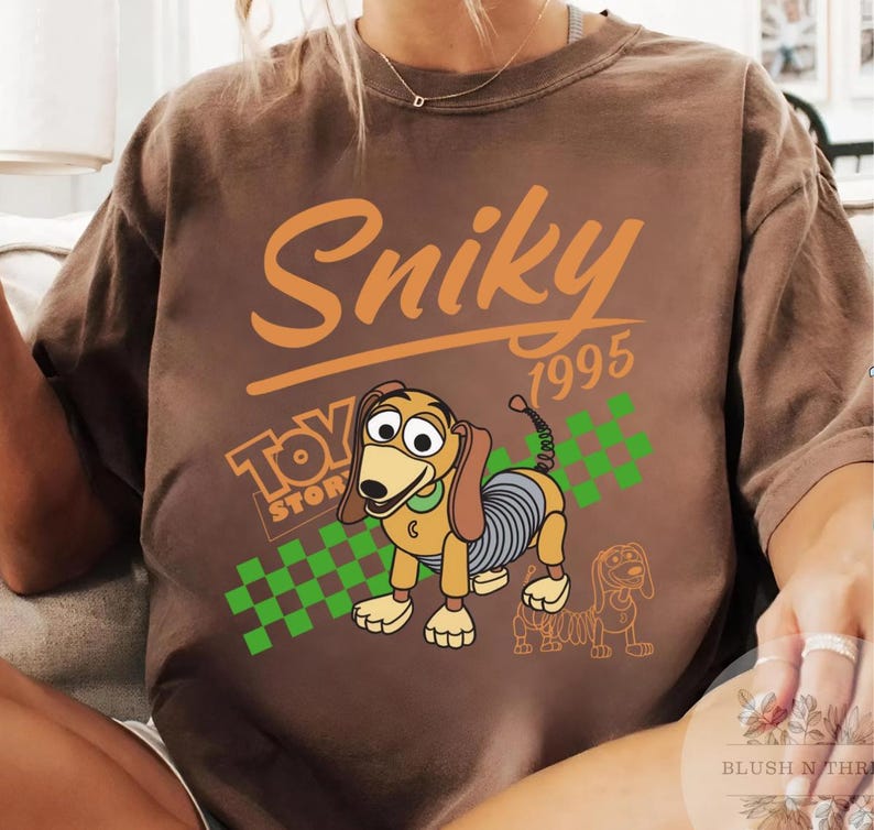 May include: Brown t-shirt featuring the text "Sniky 1995" in orange, with a cartoon image of Slinky Dog from Toy Story. The shirt also includes the Toy Story logo and a green and white checkered pattern.