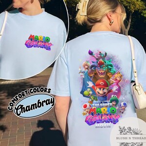 Puede incluir: Camiseta azul claro con el texto "MARIO GALAXY" en un dise&ntilde;o 3D colorido. La parte posterior de la camiseta muestra un gr&aacute;fico de personajes de la pel&iacute;cula Super Mario Galaxy. La camiseta tambi&eacute;n tiene el texto "COMFORT COLORS Chambray".
