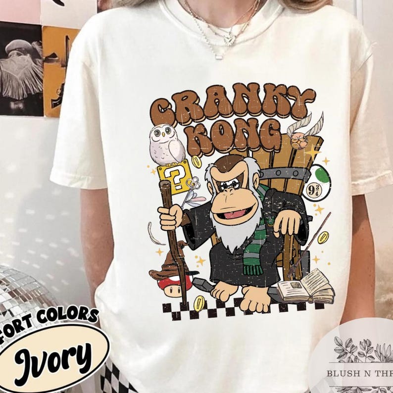 以下が含まれることがあります： アイボリー色のTシャツ。漫画のクランキーコング、フクロウ、ゲーム関連の要素のグラフィックが特徴です。「CRANKY KONG」の文字がキャラクターの上にあります。Tシャツは半袖でクルーネックです。Tシャツの色は「Ivory」です。