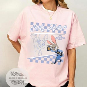 May include: A light pink t-shirt featuring a graphic of Judy Hopps from Zootopia 2. The design includes the text "Judy Hopps" in a light blue script, a checkered pattern, and an illustration of Judy Hopps in her police uniform.