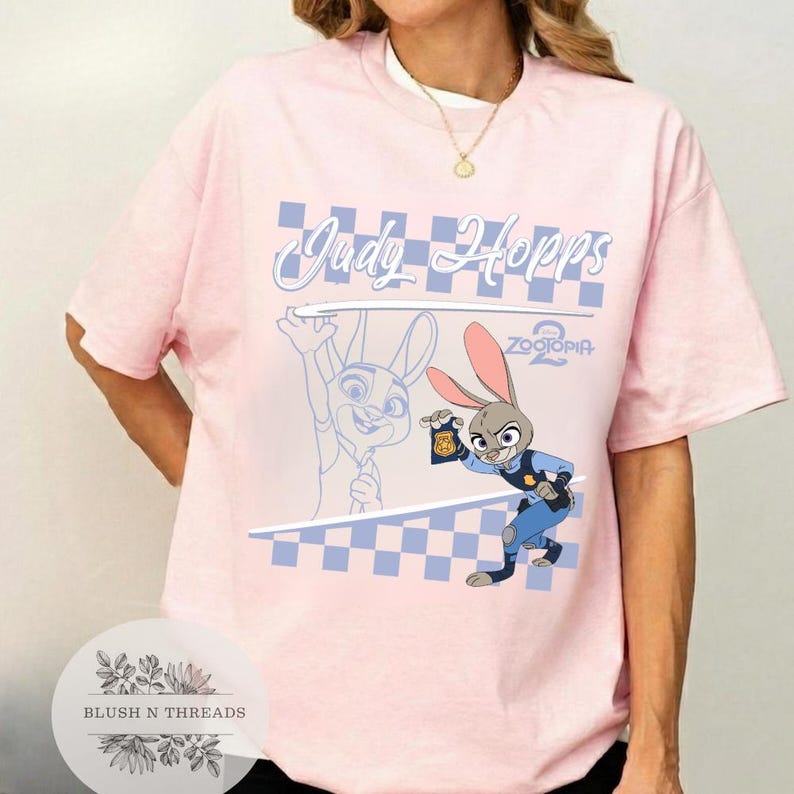 May include: A light pink t-shirt featuring a graphic of Judy Hopps from Zootopia 2. The design includes the text "Judy Hopps" in a light blue script, a checkered pattern, and an illustration of Judy Hopps in her police uniform.
