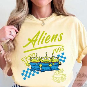 May include: A light yellow t-shirt featuring the word "Aliens" in green script above the year "1995". The shirt has a graphic of three green aliens in blue jumpsuits, with a checkered pattern and a "Toy Story" logo.