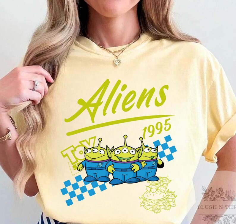May include: A light yellow t-shirt featuring the word "Aliens" in green script above the year "1995". The shirt has a graphic of three green aliens in blue jumpsuits, with a checkered pattern and a "Toy Story" logo.