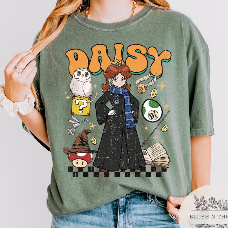 以下が含まれることがあります： プリンセスデイジーの漫画イラストが描かれた緑色のTシャツ。オレンジ色の文字で「DAISY」と書かれています。デザインにはフクロウ、クエスチョンマーク、キノコ、その他のファンタジー要素が含まれています。