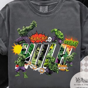 May include: Dark gray t-shirt featuring multiple illustrations of the Hulk in action poses. Comic book style with text bubbles that say "RAACK!", "BOOM!", and "WOW!". The word "HULK" is also displayed.