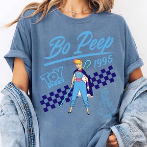 May include: A blue t-shirt featuring a graphic of Bo Peep from Toy Story. The design includes the text "Bo Peep 1995" and a checkered pattern. The character is depicted in her signature outfit, holding a staff.