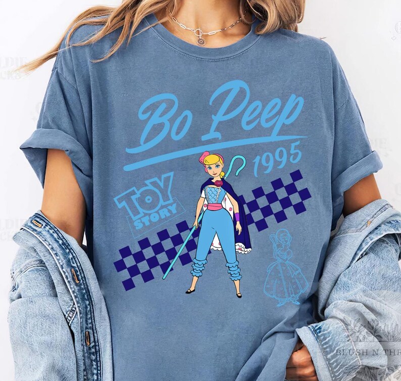 May include: A blue t-shirt featuring a graphic of Bo Peep from Toy Story. The design includes the text "Bo Peep 1995" and a checkered pattern. The character is depicted in her signature outfit, holding a staff.