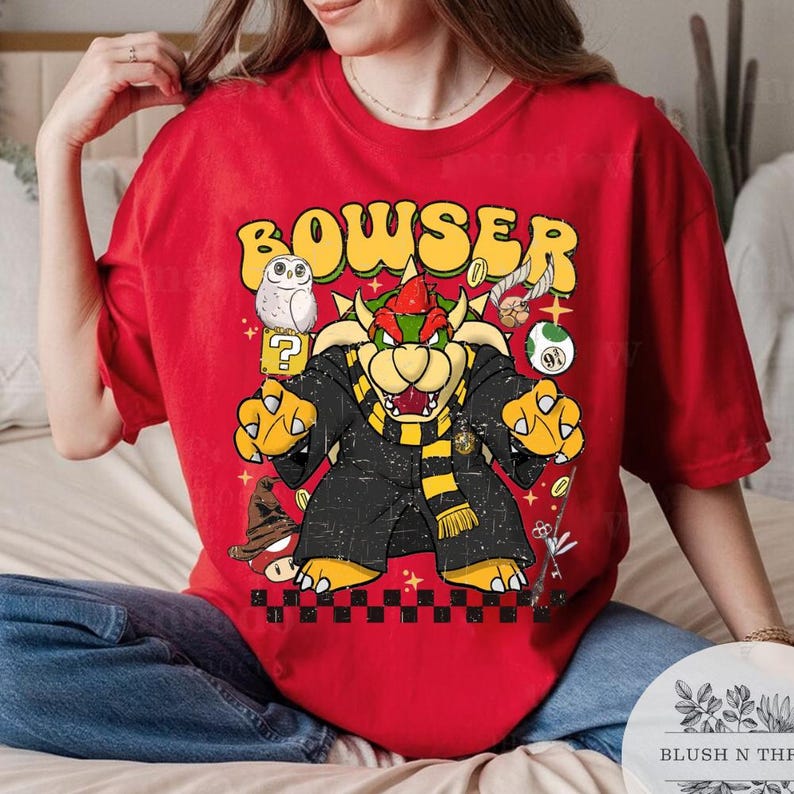 以下が含まれることがあります： 黒いローブと黄色のスカーフを着た漫画のキャラクター、クッパが描かれた赤いTシャツ。「BOWSER」の文字がキャラクターの上に黄色で書かれています。追加のグラフィックには、フクロウ、疑問符、コインが含まれています。