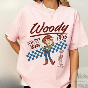 May include: A light pink t-shirt featuring the word "Woody" in a brown script font, with "1995" below. The "Toy Story" logo and a cartoon image of Woody are also present, along with a blue and white checkered pattern.