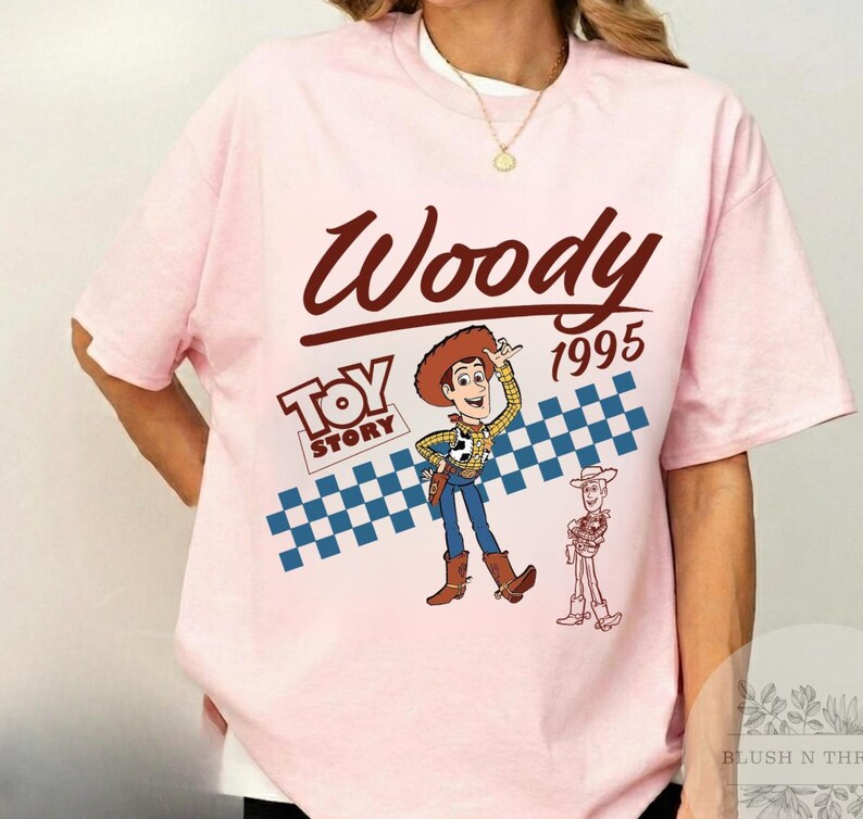 May include: A light pink t-shirt featuring the word "Woody" in a brown script font, with "1995" below. The "Toy Story" logo and a cartoon image of Woody are also present, along with a blue and white checkered pattern.