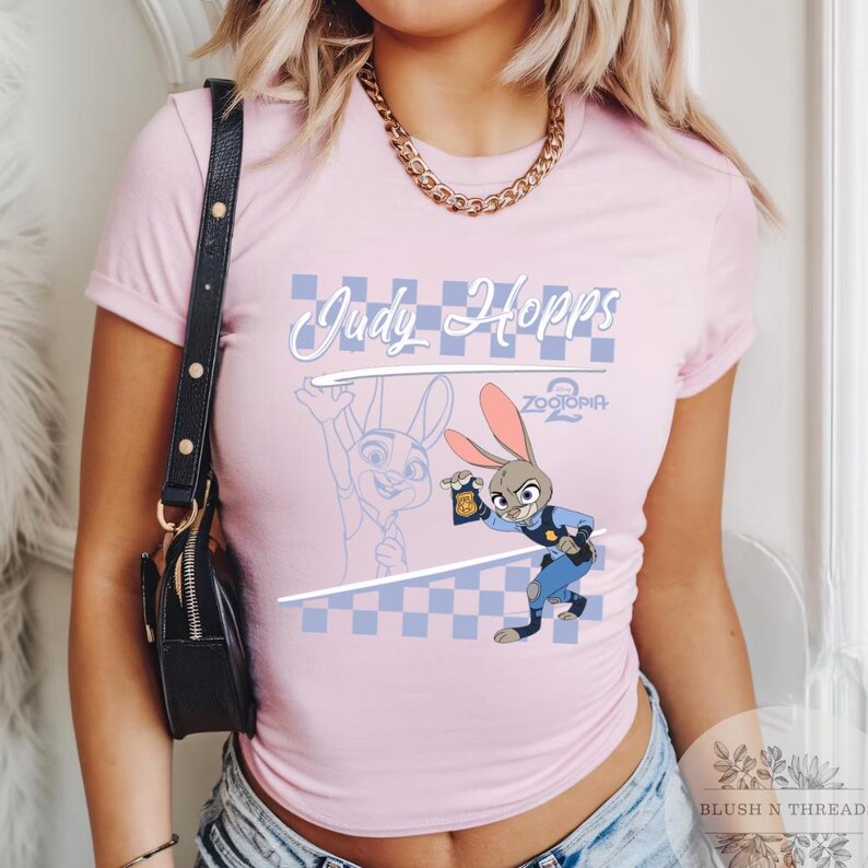 May include: A light pink t-shirt featuring the character Judy Hopps from Zootopia 2. The design includes the text "Judy Hopps" in a cursive font, a checkered pattern, and an illustration of Judy Hopps in a police uniform.
