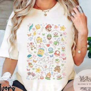 May include: Ivory-colored t-shirt featuring a colorful Super Mario design. The graphic includes various characters and elements from the game, such as Mario, Luigi, Princess Peach, and mushrooms, along with the text "Super Mario".