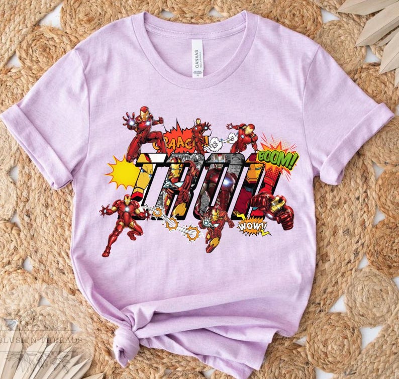 May include: A light purple t-shirt featuring a comic book style graphic of Iron Man in action poses. The word "THOR" is prominently displayed with comic book sound effects like "BOOM!" and "NOW!"