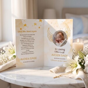 May include: A memorial card featuring gold star accents and angel wings, displaying the text Until We Meet Again, In Loving Memory of AADEN SAGE, and the dates 10.10.2025-12.12.2026. The card includes a circular photograph of a baby wrapped in a white blanket.