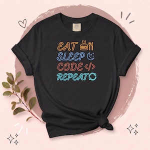 May include: A black short-sleeve t-shirt featuring a neon-style graphic with the text EAT SLEEP CODE REPEAT. The design includes icons of a burger and fork, a crescent moon with stars, code brackets, and a circular arrow symbol in orange, blue, and red.