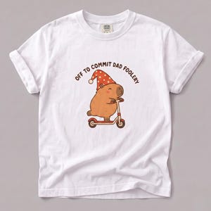 May include: A white short-sleeved t-shirt featuring a graphic of a brown capybara wearing a red polka-dot nightcap while riding a small red scooter. Above the illustration, the text reads OFF TO COMMIT DAD FOOLERY in brown capital letters.