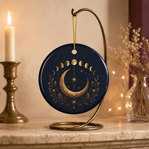 May include: A circular dark blue ceramic ornament featuring a gold-toned crescent moon, a star, and a lunar cycle phase pattern. The ornament is suspended by a thin gold cord from a curved brass-colored metal display stand.