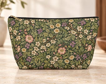 Op de afbeelding: Een tasje met ritssluiting voorzien van een dichte botanische print van roze, witte en paarse bloemen verweven met groene bladeren op een donkere achtergrond. De rechthoekige tas heeft een zwarte rits met een goudkleurige metalen trekker.