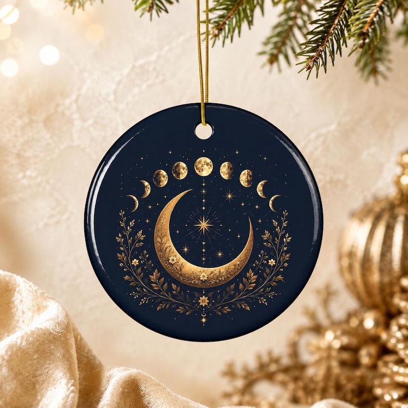 May include: A round, dark navy blue ceramic ornament featuring a gold-colored crescent moon at the center, surrounded by a lunar phase cycle, delicate floral vines, and small starburst accents. The ornament hangs from a thin gold-colored string.