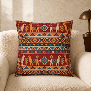 May include: A square throw pillow featuring a deep red background with repeating horizontal bands of geometric patterns and stylized llama figures in yellow, orange, blue, and white. The design includes diamond shapes and cross motifs.