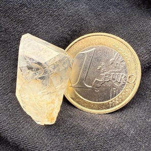 Könnte beinhalten: Ein klarer Quarz-Kristall mit goldenen Farbtönen liegt neben einer Ein-Euro-Münze. Der Kristall hat eine natürliche, ungeschliffene Form, und die Münze zeigt die Zahl 1 und das Wort "EURO". Der Hintergrund ist ein dunkles, strukturiertes Material.