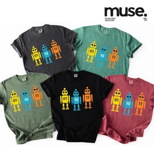 K&ouml;nnte beinhalten: Sechs kurz&auml;rmelige T-Shirts in verschiedenen Farben, jeweils mit einem bunten Roboter-Motiv in Gelb, Blau und Orange, mit den Zahlen 01, 10 und 11. Die Shirts haben den Text "muse. CUSTOM TSHIRTS LIMITED EDITIONS SINCE 2025".