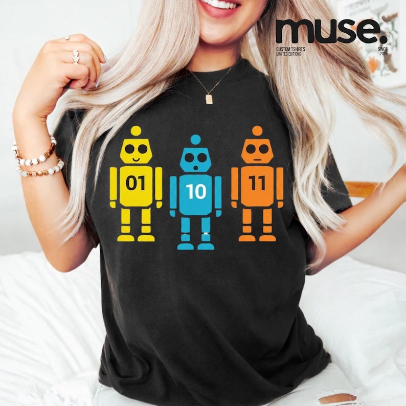 K&ouml;nnte beinhalten: Schwarzes T-Shirt mit drei bunten Roboter-Illustrationen. Die Roboter sind gelb, blau und orange, jeder mit einer anderen Nummer: 01, 10 und 11. Das Shirt hat den Text "muse" oben rechts.