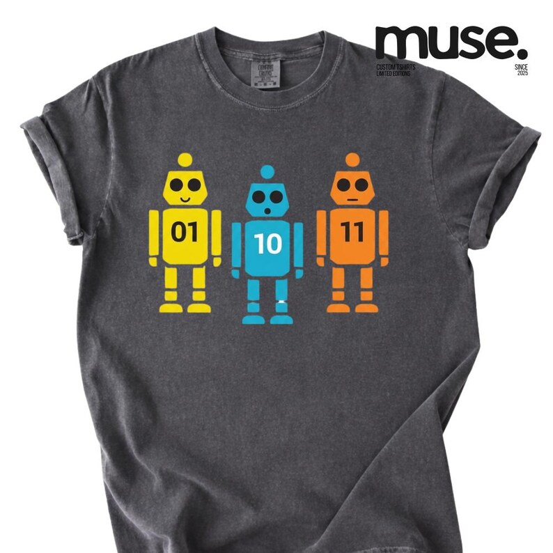 K&ouml;nnte beinhalten: Dunkelgraues T-Shirt mit drei bunten Roboter-Illustrationen. Die Roboter sind gelb, blau und orange, jeder mit einer anderen Nummer: 01, 10 und 11. Das Shirt hat hochgekrempelte &Auml;rmel und das Wort "muse" ist oben rechts aufgedruckt.