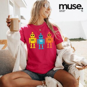K&ouml;nnte beinhalten: Ein magentafarbenes T-Shirt mit drei bunten Roboter-Designs in Gelb, Blau und Orange, jeweils mit einer Nummer: 01, 10 und 11. Der Text "muse. CUSTOM TSHIRTS LIMITED EDITIONS SINCE 2023" befindet sich oben rechts. Ein l&auml;ssiges Kleidungsst&uuml;ck.