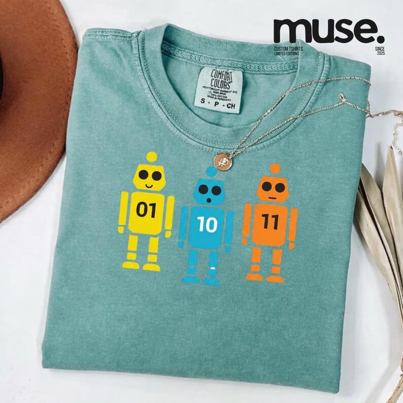 K&ouml;nnte beinhalten: Ein t&uuml;rkisfarbenes Comfort Colors T-Shirt mit drei Roboterfiguren in Gelb, Blau und Orange, jeweils mit einer Nummer: 01, 10 und 11. Das Shirt hat einen Rundhalsausschnitt und ein kleines Etikett. Eine silberne Halskette mit Anh&auml;nger liegt auf dem Shirt.