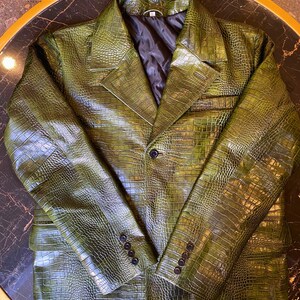 May include: A dark olive green blazer featuring a textured crocodile skin pattern. The jacket has a notched lapel, a single front button closure, and four small black buttons on each sleeve cuff. It includes a black quilted interior lining.