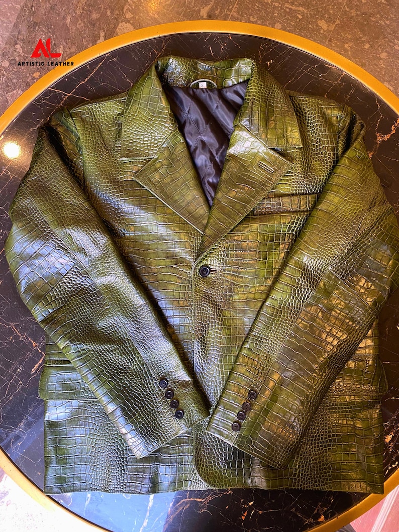 May include: A dark olive green blazer featuring a textured crocodile skin pattern. The jacket has a notched lapel, a single front button closure, and four small black buttons on each sleeve cuff. It includes a black quilted interior lining.