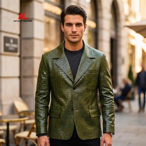 May include: An olive green leather blazer with a textured crocodile skin pattern, featuring a notched lapel, two front buttons, and two flap pockets. The upper left corner displays the red and black text logo for Artistic Leather Studio.