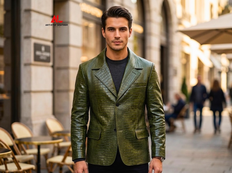 May include: An olive green leather blazer with a textured crocodile skin pattern, featuring a notched lapel, two front buttons, and two flap pockets. The upper left corner displays the red and black text logo for Artistic Leather Studio.