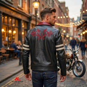 Könnte beinhalten: Eine schwarze Lederjacke mit dunkelrotem Kragen, einem roten, gestickten Tribal-Design auf dem oberen Rücken und zwei horizontalen cremefarbenen Streifen an jedem Ärmel. Die Jacke wird mit einer dunkelblauen Jeans im Freien getragen.