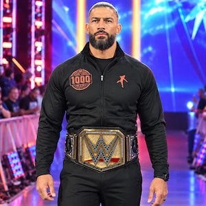 May include: A black zip-up track jacket featuring an orange graphic of a person jumping on the left chest and an orange circular emblem with the number 1000 on the right chest, worn with a large gold and black championship belt around the waist.