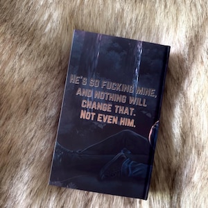 K&ouml;nnte beinhalten: Ein Buch mit dunklem Einband mit dem Text "HE'S SO FUCKING MINE, AND NOTHING WILL CHANGE THAT. NOT EVEN HIM." Das Cover zeigt eine dunkle Waldszene mit einer liegenden Person.