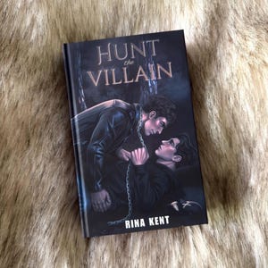 K&ouml;nnte beinhalten: Ein Buch mit dem Titel "Hunt the Villain" von Rina Kent. Das Cover zeigt zwei M&auml;nner in einer dramatischen Szene mit einer Kette. Der Titel ist in einer stilisierten Schriftart. Das Buch liegt auf einem Kunstfell-Hintergrund.