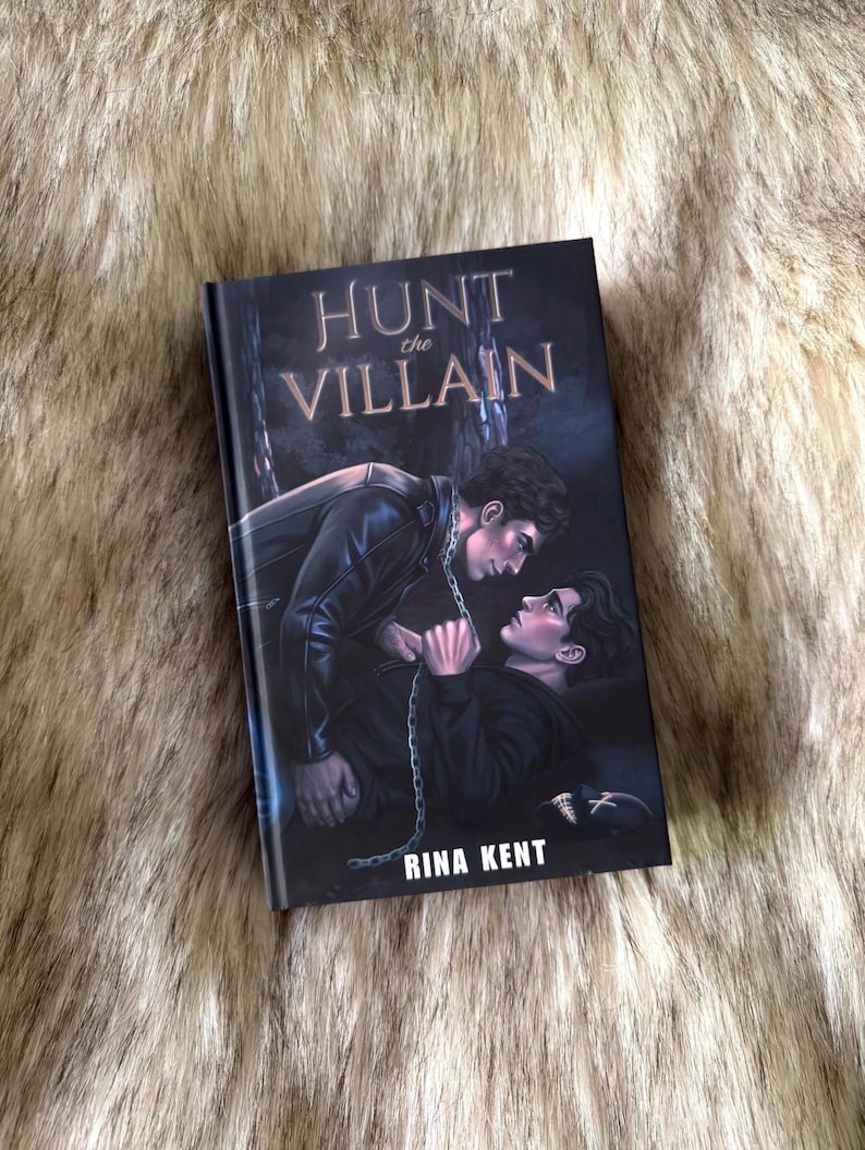 K&ouml;nnte beinhalten: Ein Buch mit dem Titel "Hunt the Villain" von Rina Kent. Das Cover zeigt zwei M&auml;nner in einer dramatischen Szene mit einer Kette. Der Titel ist in einer stilisierten Schriftart. Das Buch liegt auf einem Kunstfell-Hintergrund.