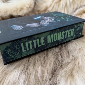Pu&ograve; includere: Un libro con copertina rigida con una copertina scura con l'illustrazione di due figure. Il dorso del libro mostra il testo "LITTLE MONSTER" in lettere verdi. Il libro &egrave; appoggiato su una superficie soffice e chiara.
